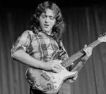 Rory Gallagher in a black and white photo, head tilted, holding his iconic Stratocaster guitar, wearing a checked shirt.